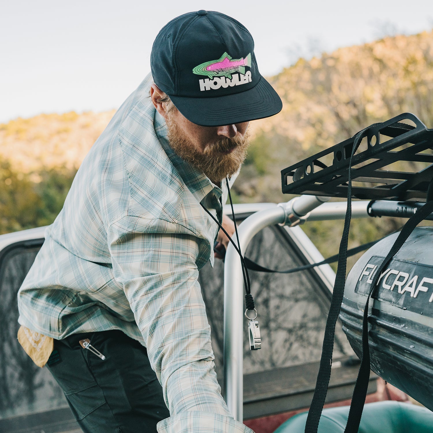 A bearded man in a plaid shirt and cap with a soft crown loads gear onto a vehicle roof rack beside an inflatable boat labeled FLYCRAFT in an outdoor, wooded setting. - View 2