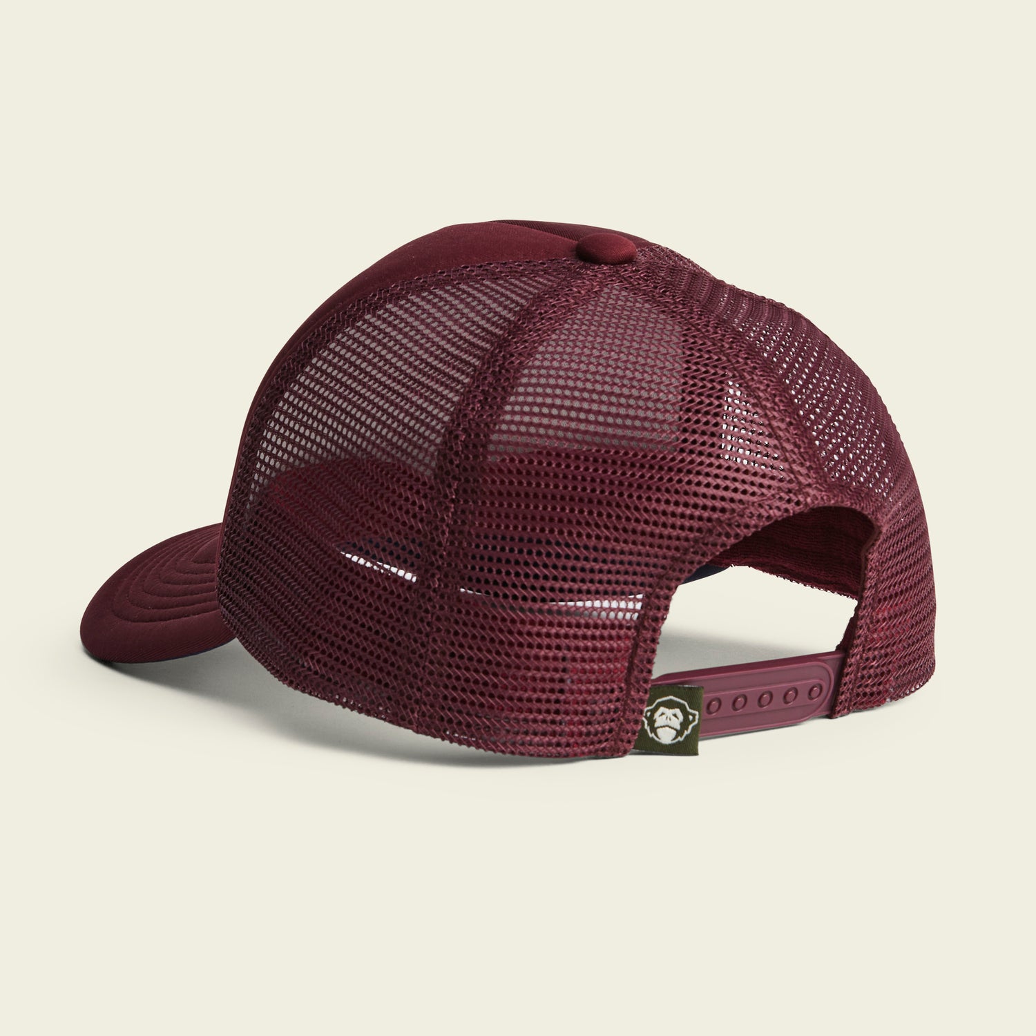 A maroon mesh trucker hat shown from the back, featuring an adjustable snapback closure and a small green tag with a cartoon monkey face logo near the strap—this vintage-inspired hat boasts classic retro style. - View 3