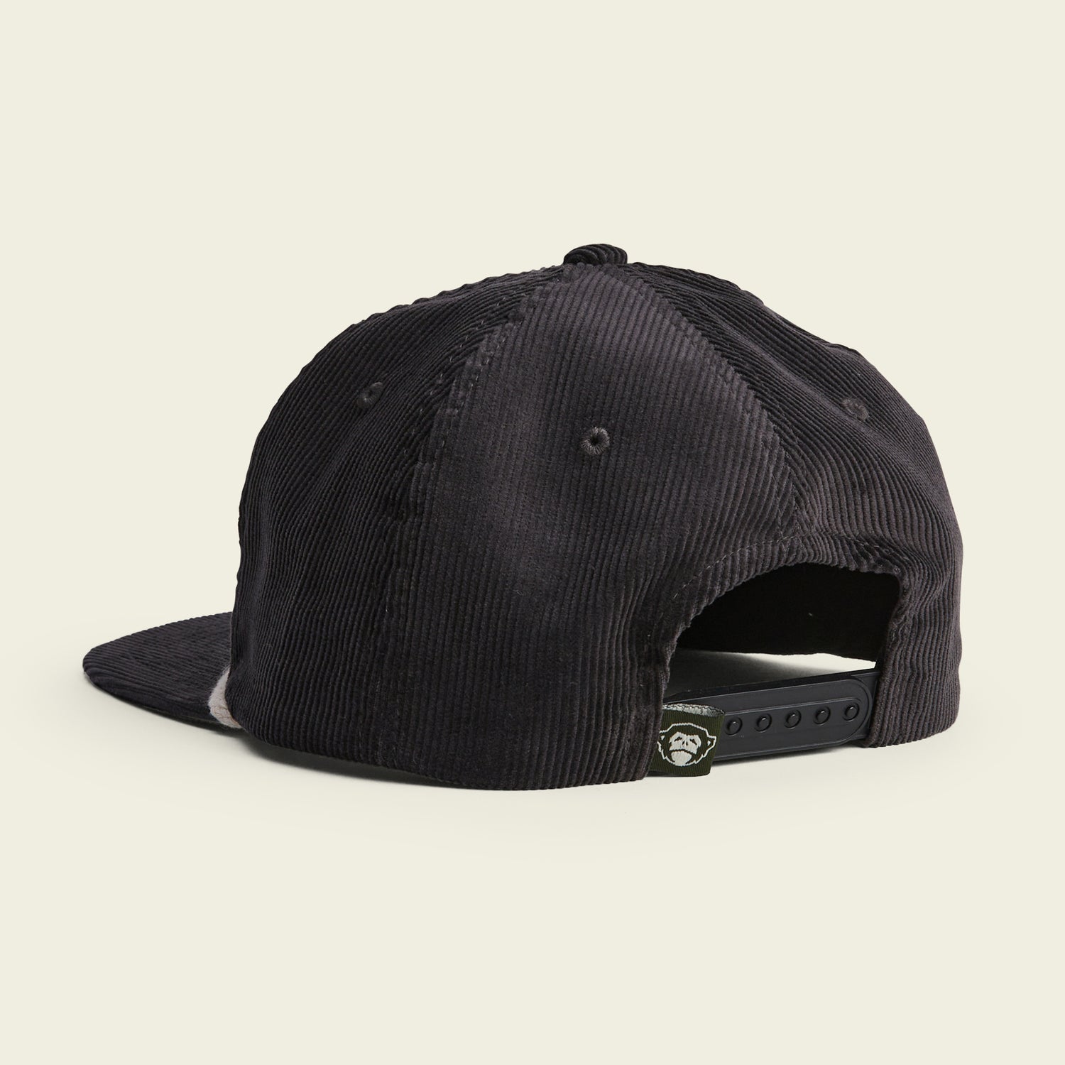 A black corduroy unstructured snapback hat shown from the back, featuring a soft crown, adjustable snapback closure, and a small logo tag near the opening. The background is a plain, light beige color. - View 3
