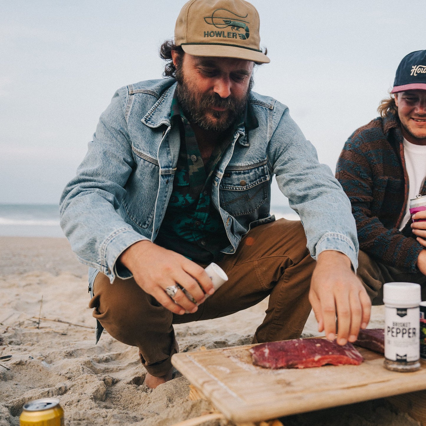 A man in a denim jacket kneels on a sandy beach, seasoning raw meat on a wooden board. Another casually dressed person sits nearby, with the ocean and sky stretching in the background. - View 2