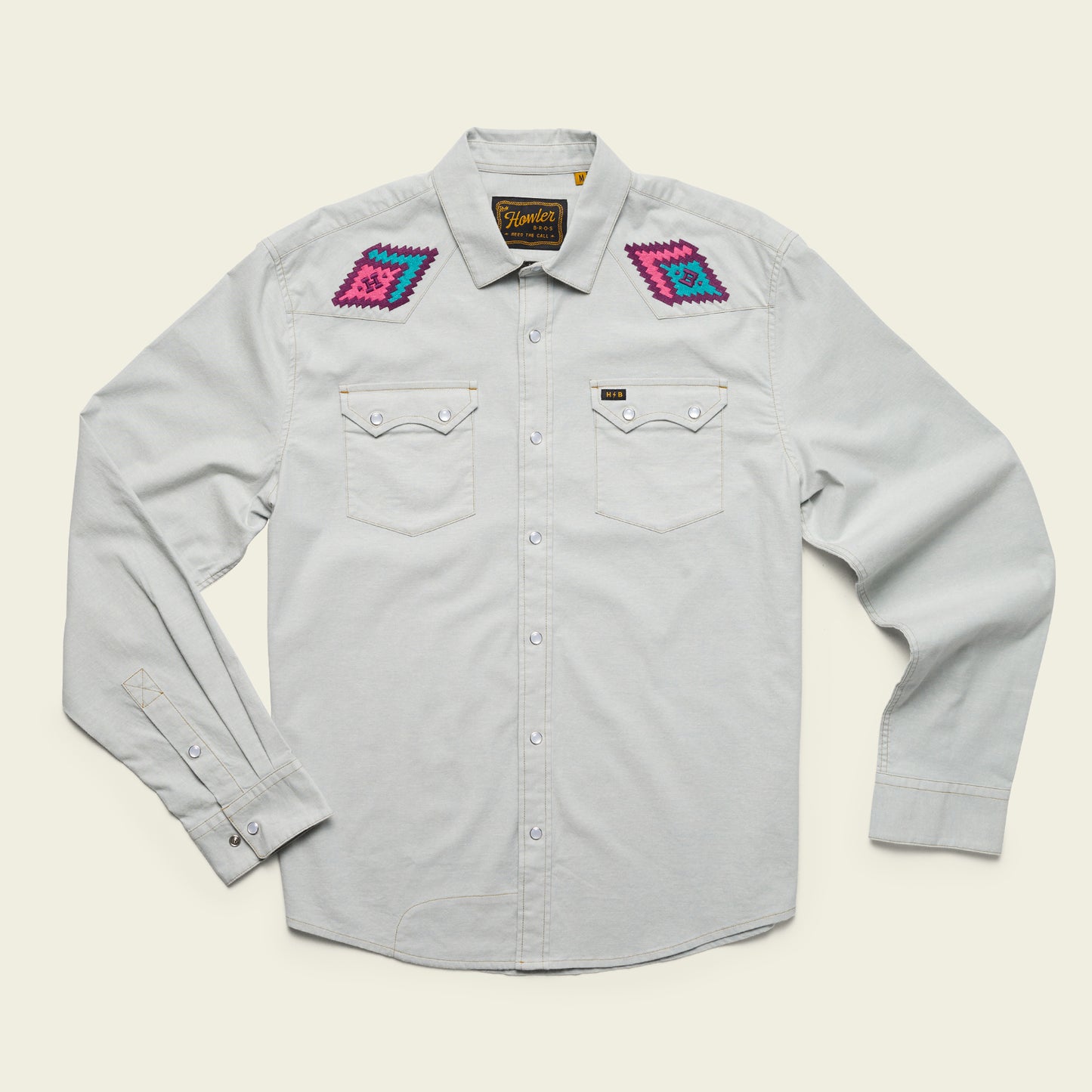 A light gray, long-sleeve stretch chambray button-up shirt with pearl snaps, two chest pockets, and colorful, vintage inspired motif embroidery in pink, purple, and blue on each shoulder. The shirt is laid flat on a cream background. - View 11