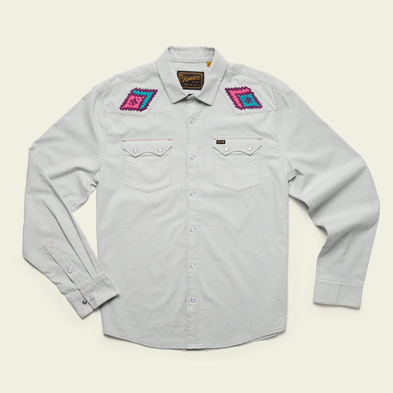 A light gray, long-sleeve stretch chambray button-up shirt with pearl snaps, two chest pockets, and colorful, vintage inspired motif embroidery in pink, purple, and blue on each shoulder. The shirt is laid flat on a cream background. - View 11