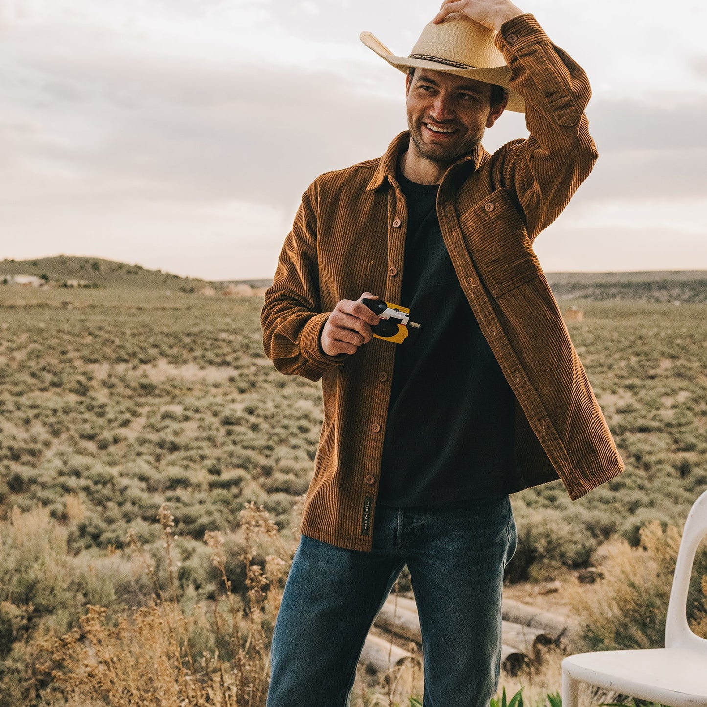 A smiling man in a brown jacket and jeans stands outdoors in a field, holding a small toy and adjusting his cowboy hat with one hand. His Iquitos Overshirt adds rugged style against the open countryside and cloudy sky. - View 2