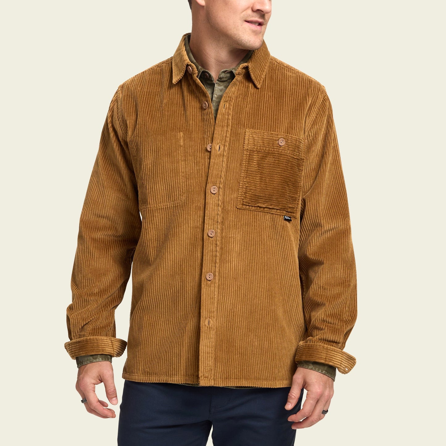 A man wearing a tan wide wale corduroy Iquitos Overshirt with a front chest pocket, layered over a green shirt and dark pants. He stands with his head turned slightly to the side—a perfect travel shirt look. - View 4
