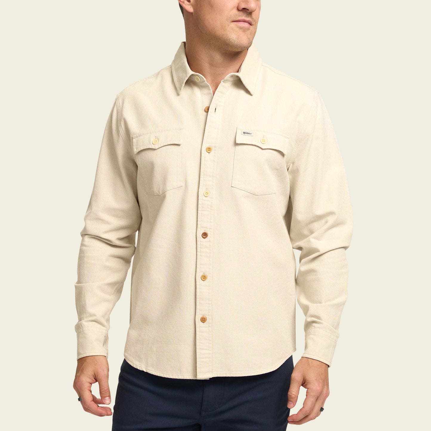 A man wearing a long-sleeve, cream-colored Novato Shirt with two chest pockets and dark pants stands against a plain light background. - View 4