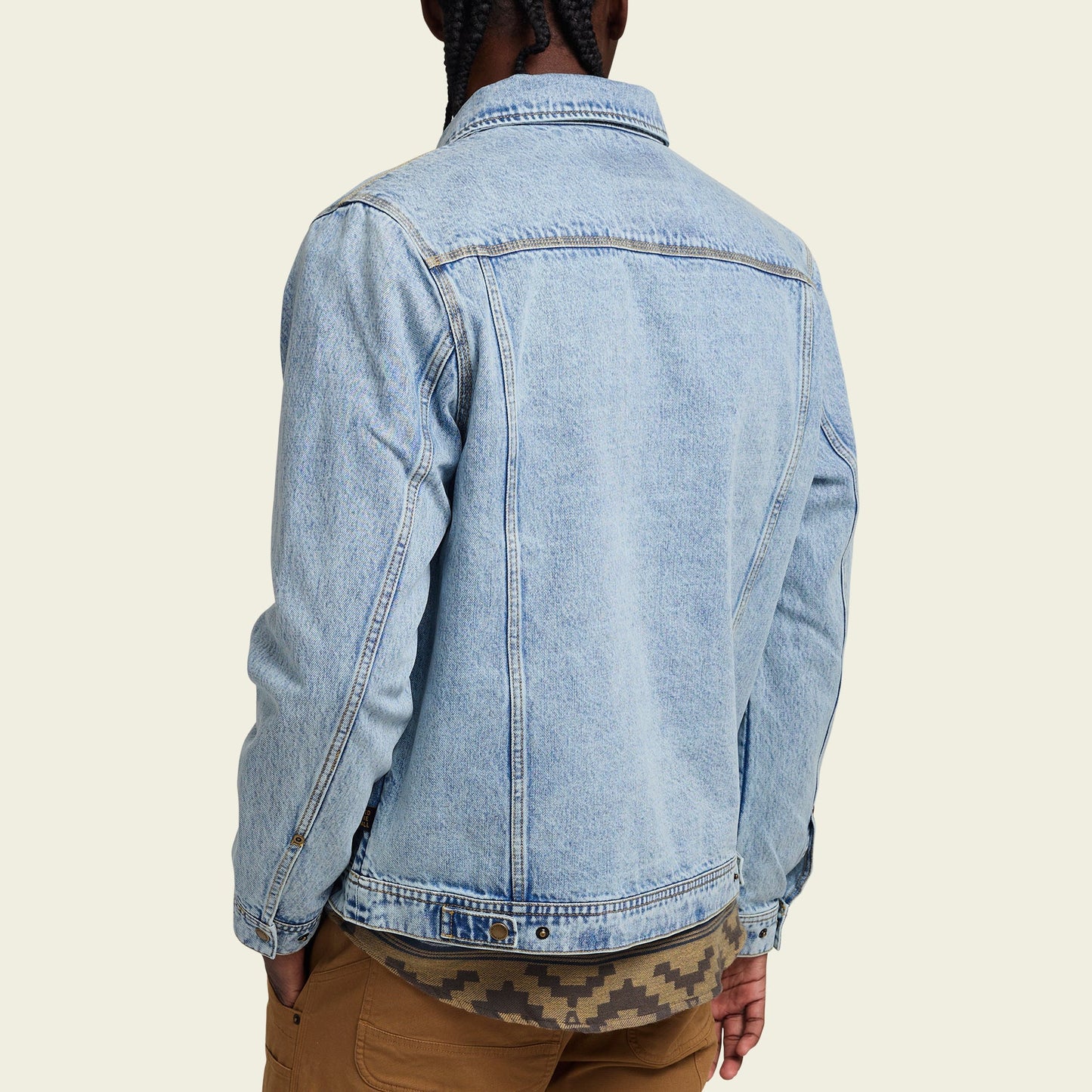 A person with braided hair is shown from the back, wearing a light blue denim jacket with a distinctive lightning bolt stitch over a patterned shirt and brown pants against a light beige background. - View 6