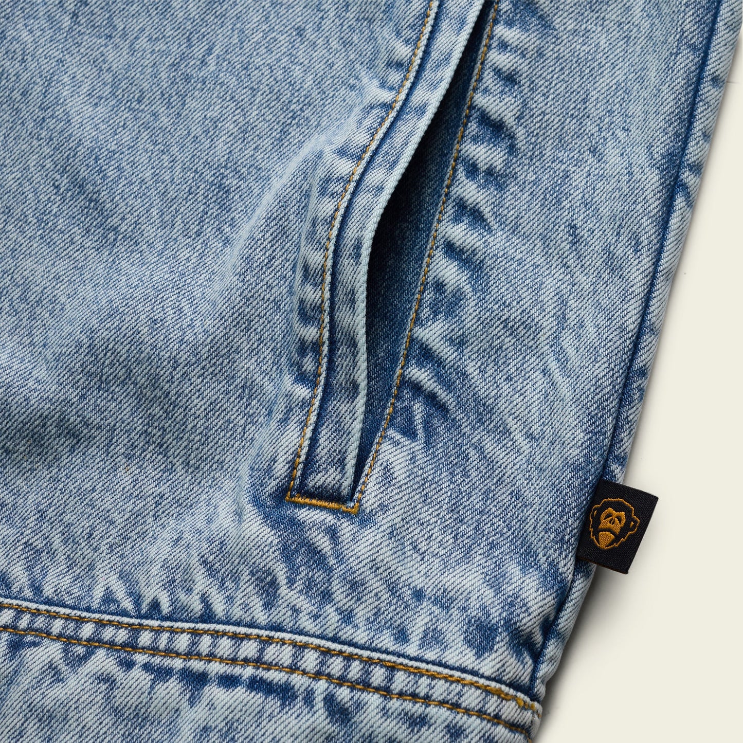 Close-up of light blue denim fabric, likely from a Depot Jacket, showing a vertical pocket with yellow lightning bolt stitch and a small black tag featuring a gold ape face logo on the seam. - View 8