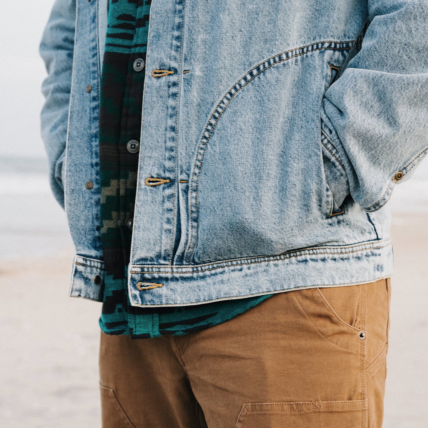 A person stands on the beach with hands in the pockets of a light blue Depot Jacket featuring a subtle lightning bolt stitch, wearing a green patterned shirt underneath and tan pants. The ocean and sand are visible in the blurred background. - View 3