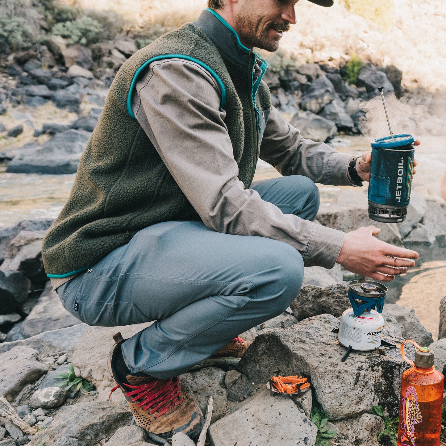 A person in outdoor gear and tech fabric pants kneels on rocks by a river, warming their hands over a portable camping stove with a blue Jetboil cooking system. An orange fuel canister and multitool are nearby. - View 2