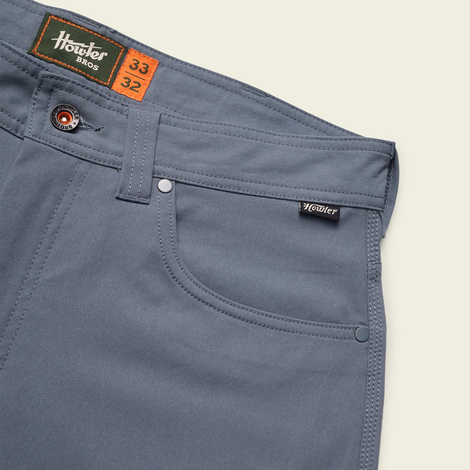 Close-up of blue Howler Bros Frontside Tech Pants showing front pocket, belt loops, rivets, and branded labels. These 5-pocket pants feature size tags indicating waist 33 and inseam 32. - View 6