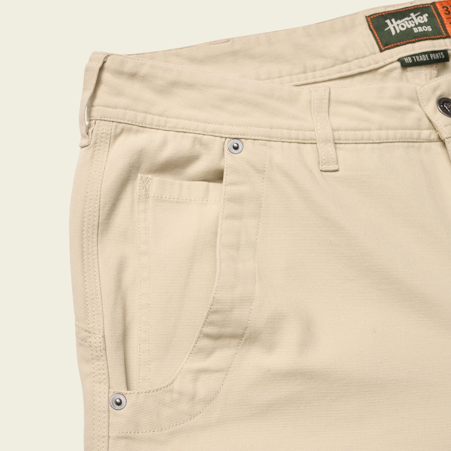 Close-up of the front pocket and waistband of light beige utility pants with visible stitching, silver rivets, and a green and orange “Howler Bros” label on the inside waistband. - View 6