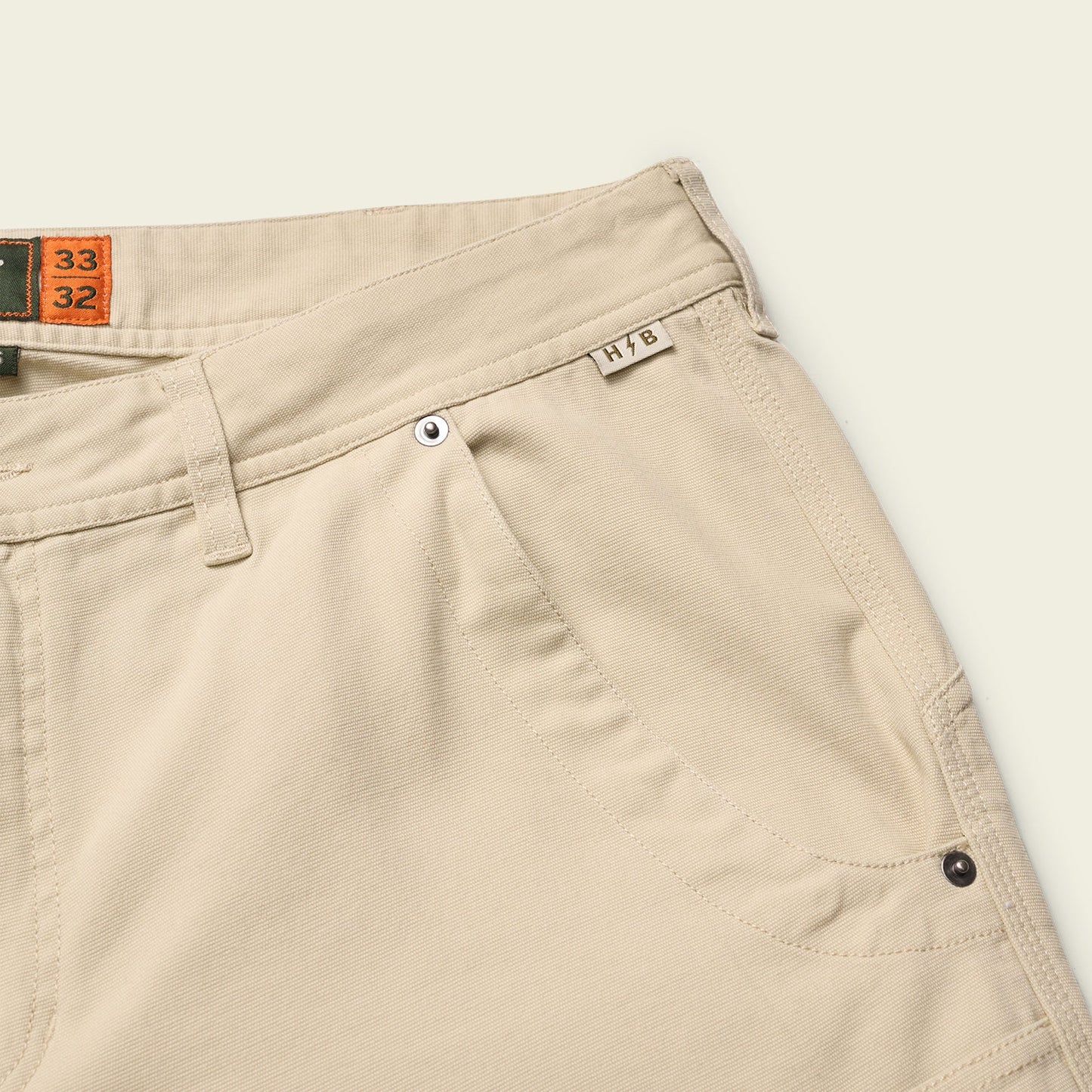 Close-up of beige workwear pants showing the waistband, belt loops, front pocket with metal rivets, and a small fabric tag labeled HTB. Size tags 33 and 32 are visible inside the waistband. - View 3