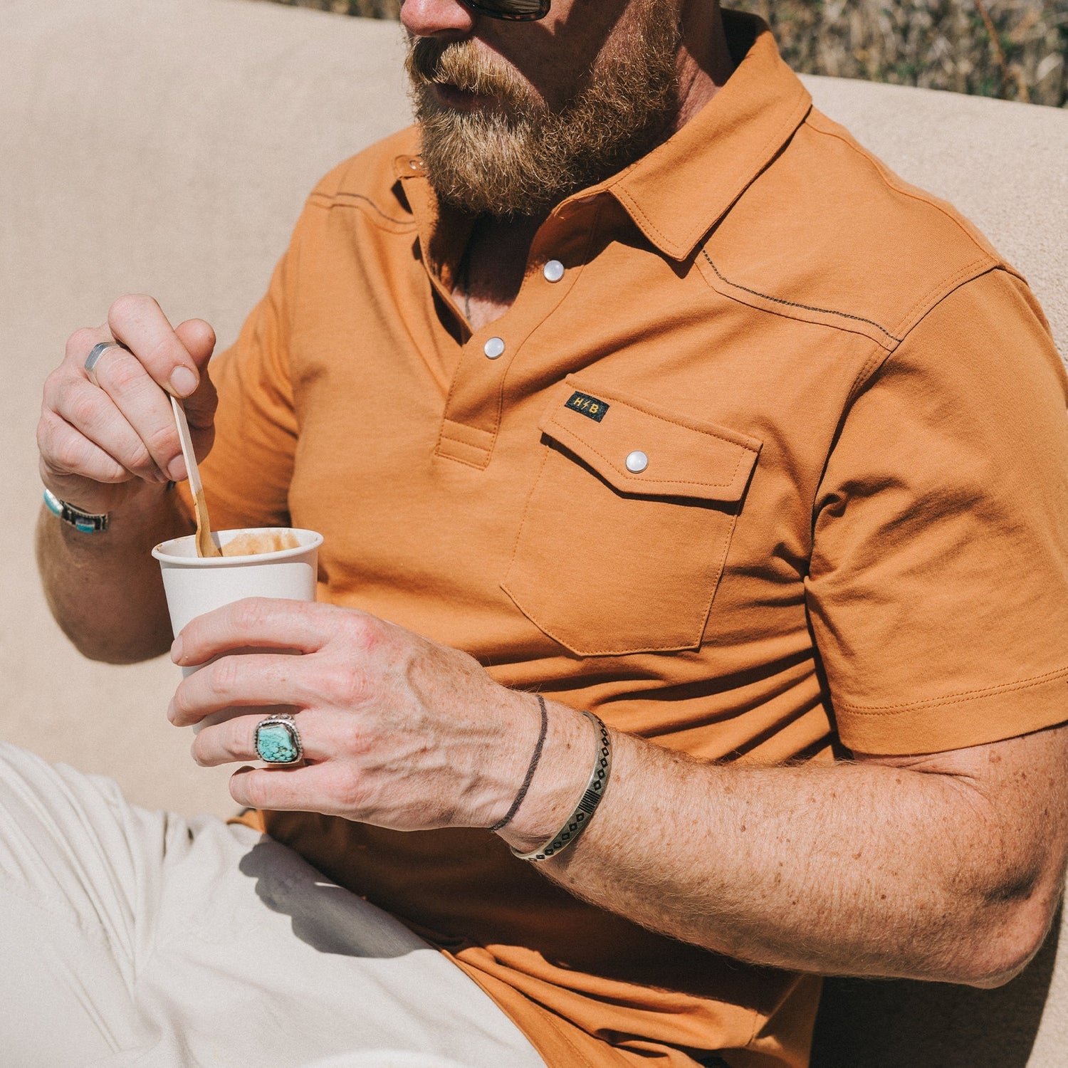 A man in a heathered cotton Ranchero Polo and sunglasses holds a cup, stirring with a spoon. He has a beard, wears rings, bracelets including a turquoise ring, and sits outdoors in the sunlight. - View 2