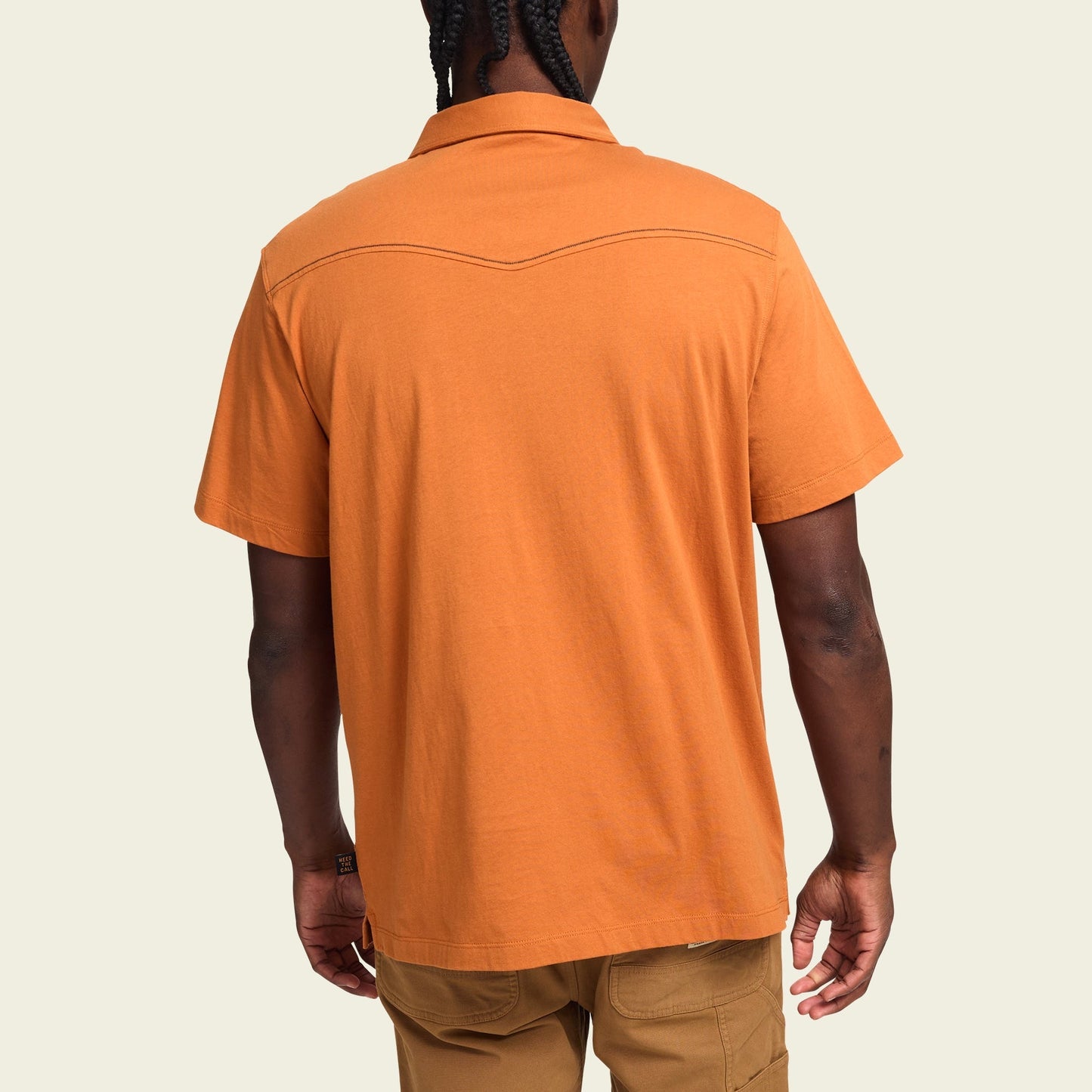 A person with braided hair is shown from the back, wearing an orange western style polo short-sleeve collared shirt crafted in heathered cotton and light brown pants, set against a plain light background. - View 5
