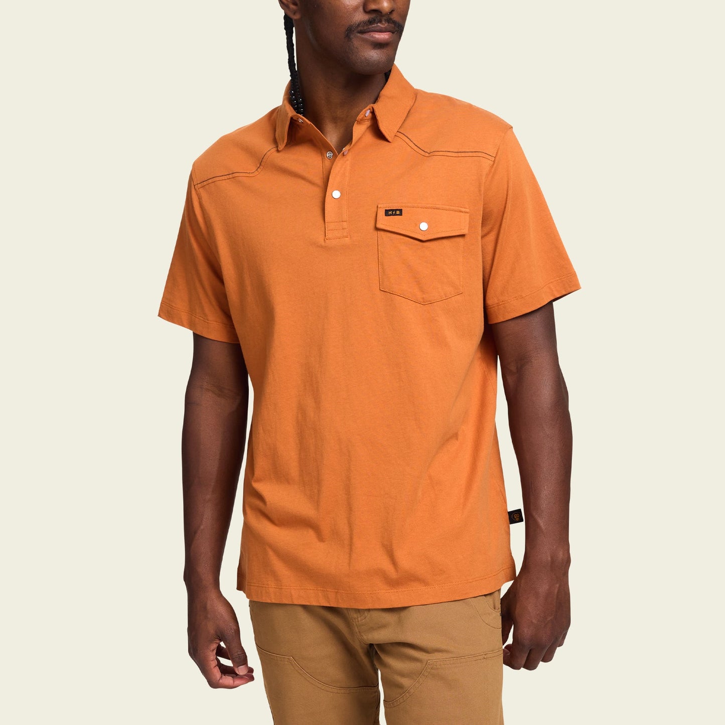 A man wearing an orange heathered cotton Ranchero Polo with a buttoned chest pocket and tan pants, standing against a plain light background. - View 4