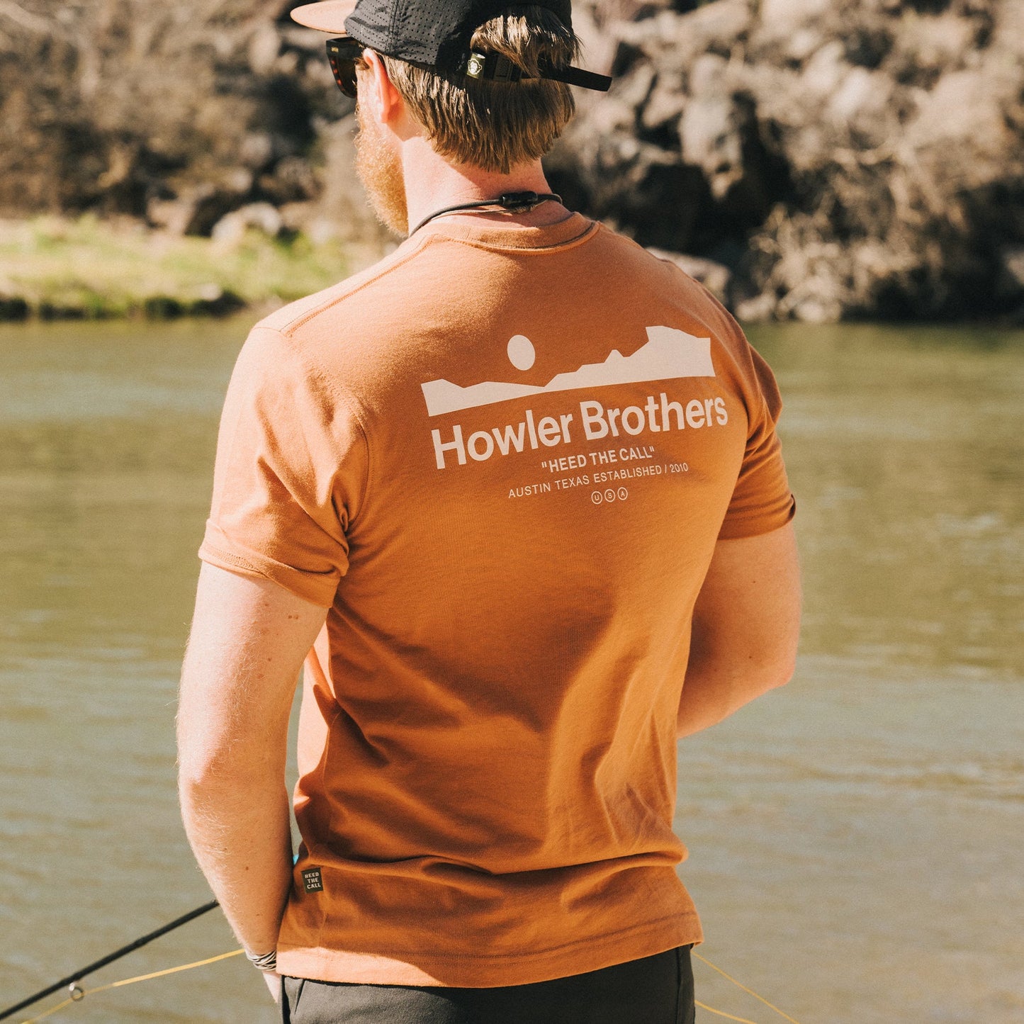 A man wearing an orange Howler blended t-shirt stands by a river, holding fishing gear, with rocky terrain and water in the background, enjoying maximum comfort from its cotton-poly blend. - View 2