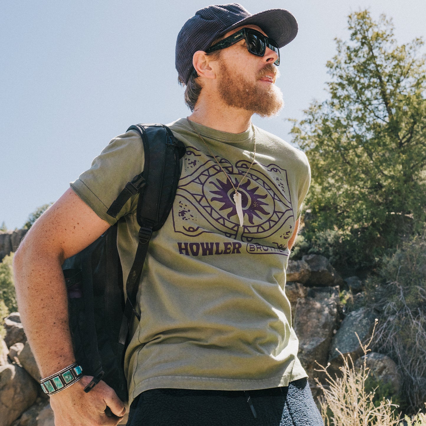 A bearded man wearing sunglasses, a cap, and an olive graphic T-shirt with HOWLER on it stands outdoors with a black backpack, evoking the laid-back style of vintage t-shirts. Trees and rocks are visible in the background. - View 2