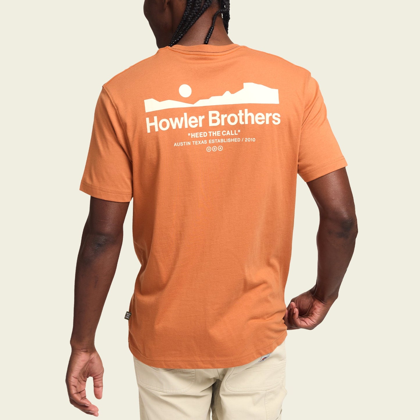 A person wearing an orange Howler blended t-shirt with white text and a landscape graphic on the back. Made from a cotton-poly blend for maximum comfort, the shirt reads “Howler Brothers ‘Heed the Call’ Austin, Texas Established 2010.” The person is facing away. - View 4