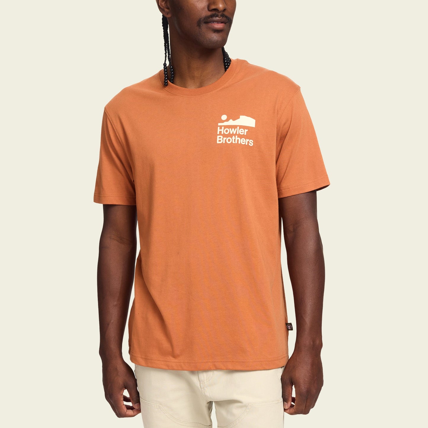 A man wearing a burnt orange Howler blended t-shirt with a small white Howler Brothers logo on the left chest, standing against a plain light background. The cotton-poly blend ensures maximum comfort, paired with light-colored pants. - View 3