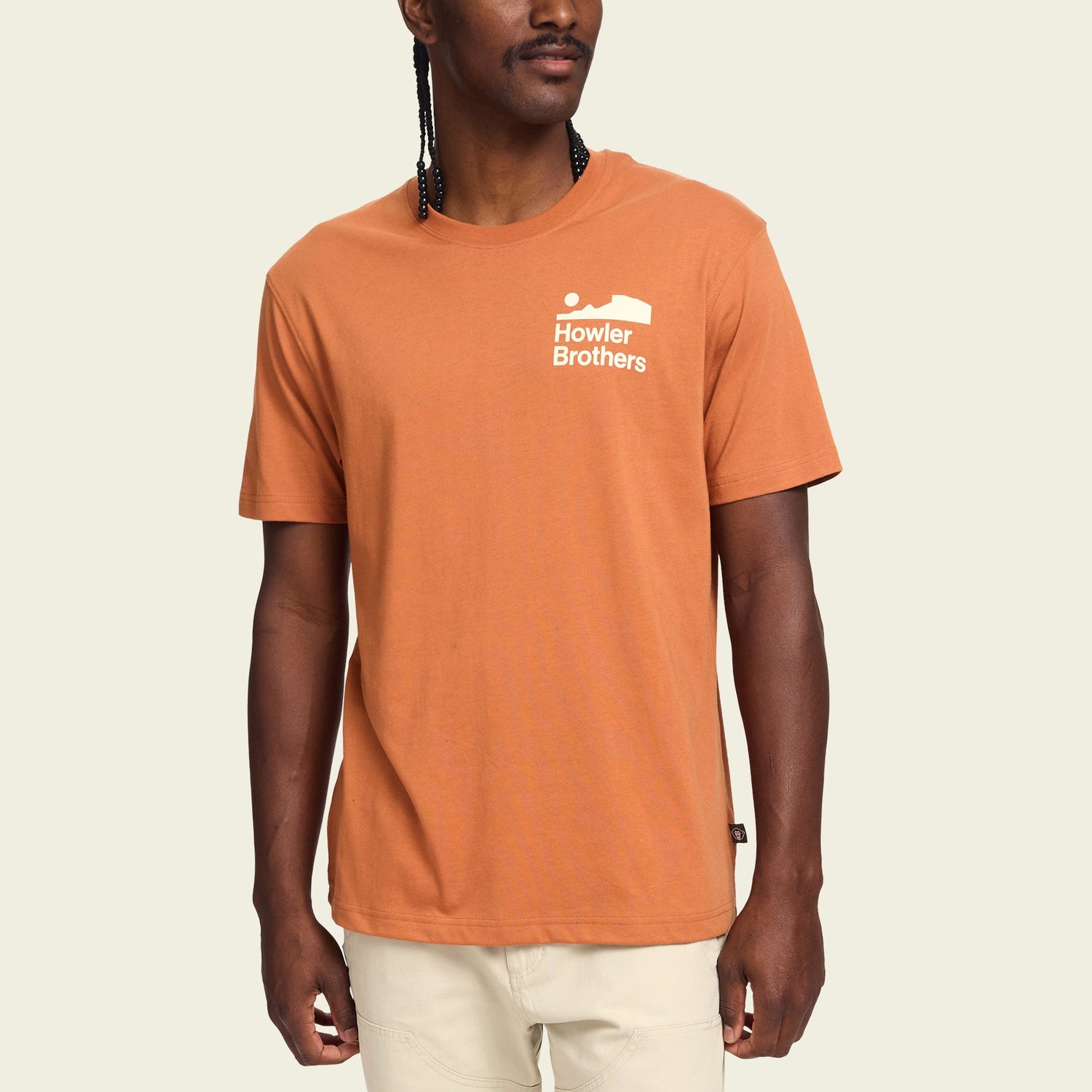 A man wearing a burnt orange Howler blended t-shirt with a small white Howler Brothers logo on the left chest, standing against a plain light background. The cotton-poly blend ensures maximum comfort, paired with light-colored pants. - View 3
