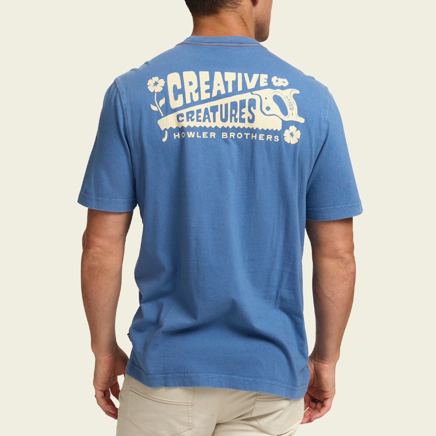 A person wearing a blue short-sleeve t-shirt with Creative Creatures and Howler Brothers printed on the back in white, along with a hand saw graphic and flower illustrations, evokes the spirit of vintage t-shirts. The person is facing away from the camera. - View 4