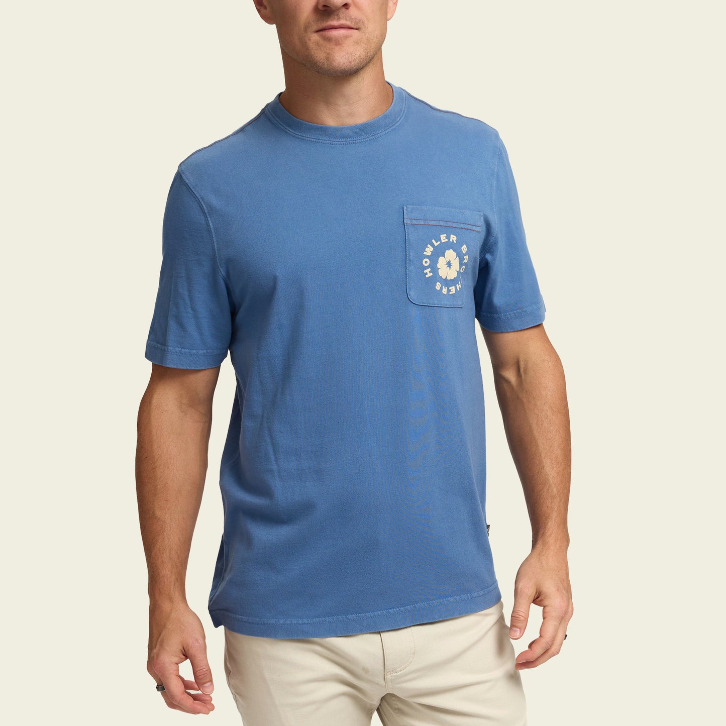 A man wearing a blue short-sleeve t-shirt with a chest pocket featuring experimental artwork and text, paired with light-colored pants, stands against a plain light background. - View 3
