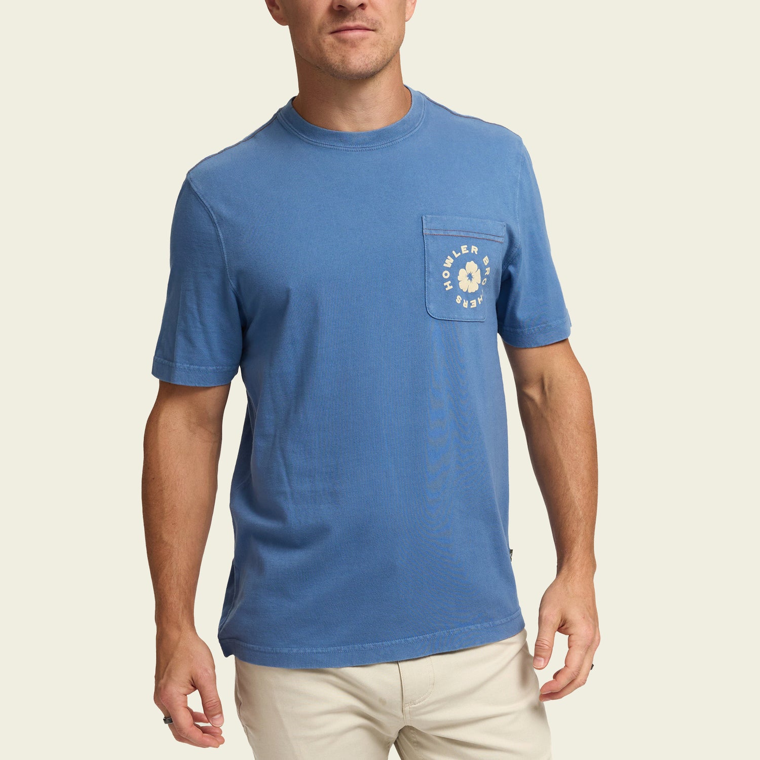 A man wearing a blue short-sleeve t-shirt with a chest pocket featuring experimental artwork and text, paired with light-colored pants, stands against a plain light background. - View 3