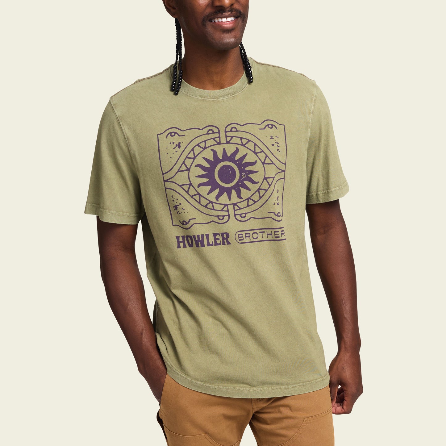 A person wearing a light olive green T-shirt with a graphic of two stylized alligators and a sun in the center, blending experimental artwork with the vibe of vintage t-shirts. The text reads “HOWLER BROTHER.” The person smiles, one hand in their pocket. - View 3
