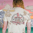 A person with curly hair faces away, wearing a tie-dye cotton t-shirt featuring a wizard holding a fish with Howler Brothers and Heed the Call text. This vintage look is accented by cartoon mushrooms, sparkles, a lake, and mountains.