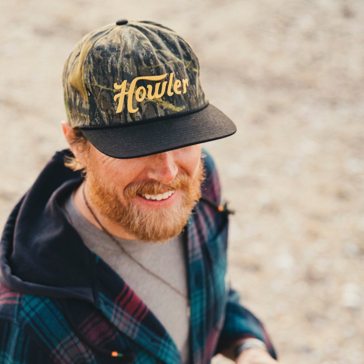 A smiling man with a reddish beard wears a camouflage Howler cap featuring a soft crown and adjustable snapback closure, along with a plaid jacket, standing outdoors on a blurred, sandy background. - View 4