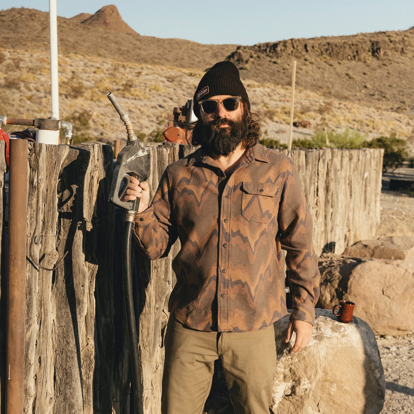A bearded man wearing sunglasses, a beanie, and a buttery soft flannel stands outdoors by a rustic wooden fence, holding a metal kettle in one hand. Desert mountains and dry vegetation are visible in the background. - View 2