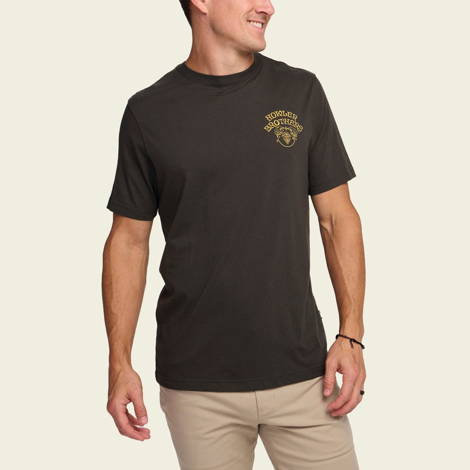 A man wearing a black Howler t-shirt with a small yellow logo on the left chest stands smiling in light khaki pants against an off-white background, showing off the comfort of these cotton-poly blend comfortable t-shirts. - View 4