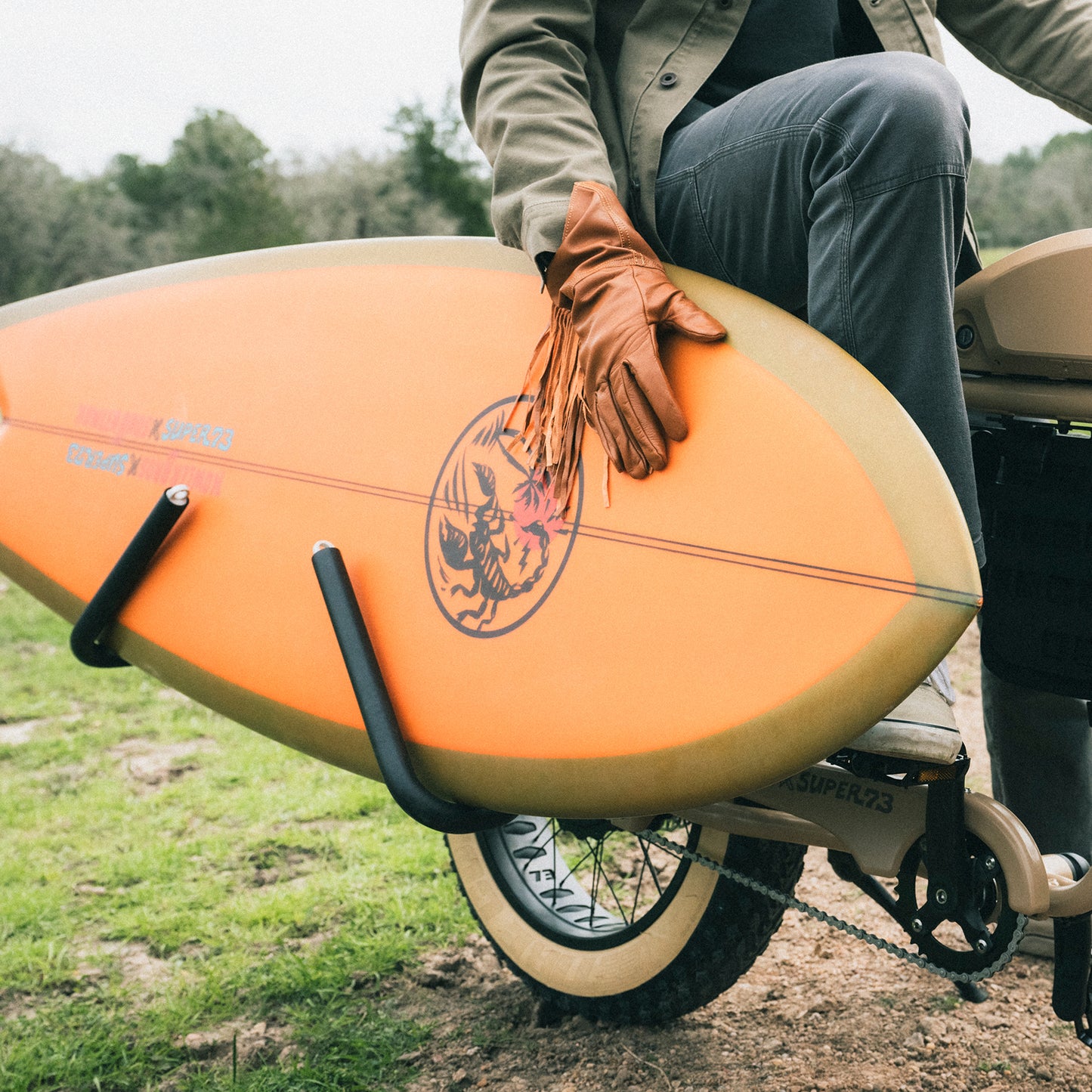 A person wearing brown leather gloves holds an Almond Surfboards orange surfboard while sitting on a Super73-S2 bicycle with surfboard racks, on a grassy outdoor path. - View 2