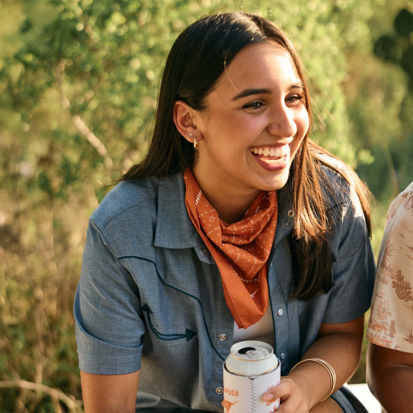 A young woman with long brown hair, wearing a blue shirt and a Los Longhorns de Tejas Bandana, sits outdoors smiling and holding a can. Sunlight and greenery are in the background, perfect for showing off her game day accessories. - View 2