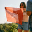 A woman in sunglasses and denim shorts smiles outdoors, holding up a Los Longhorns de Tejas Bandana—one of the must-have game day accessories—against a backdrop of green trees on a sunny day.