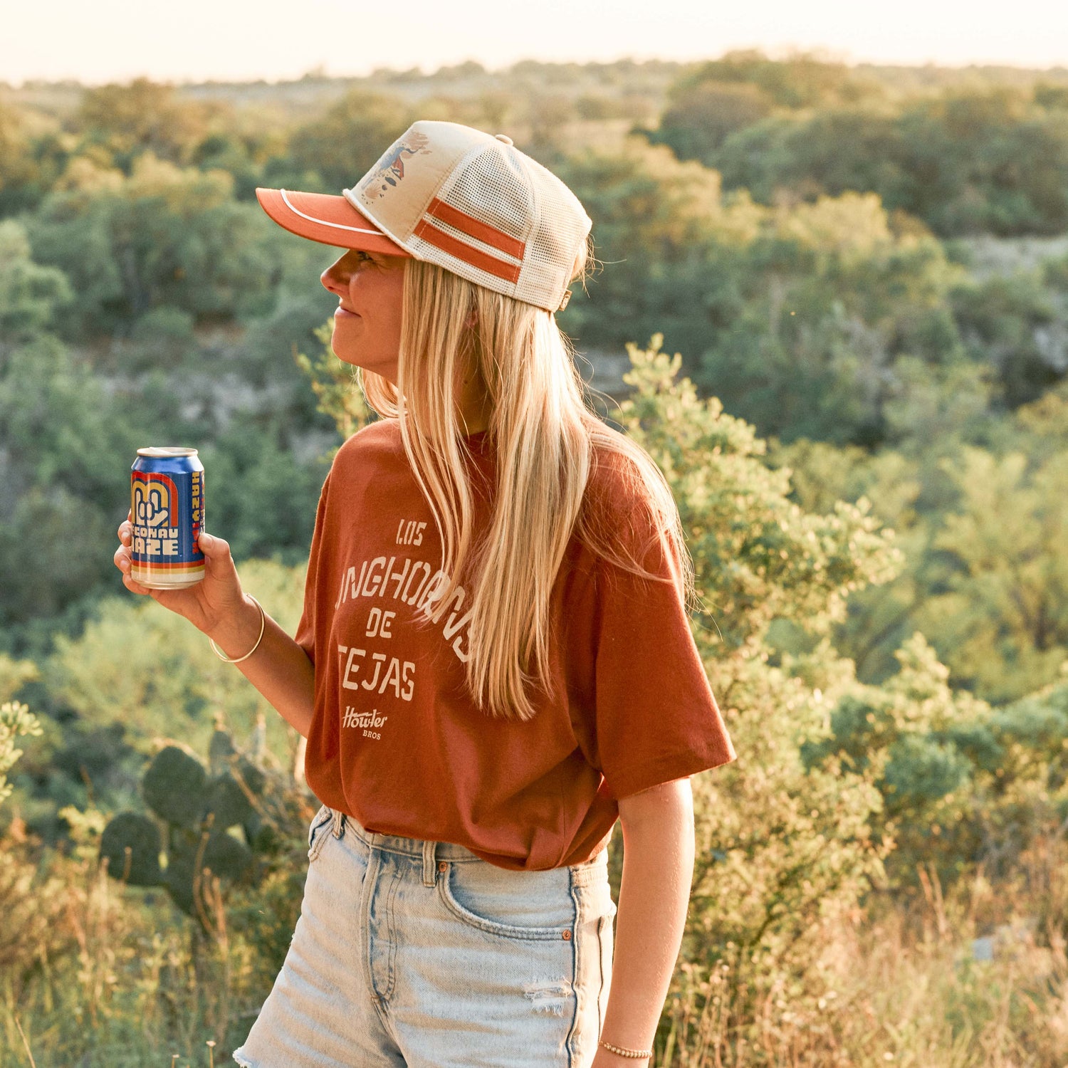 A woman in a red T-shirt and denim shorts stands outdoors, holding a can of soda. She sports sunglasses and a vintage-inspired hat, with greenery and hills bathed in warm sunlight as her scenic backdrop. - View 3