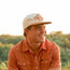 Smiling young man wearing a patterned orange shirt and an Unstructured Snapbacks cap, sitting outdoors with trees and a bright sky in the background.