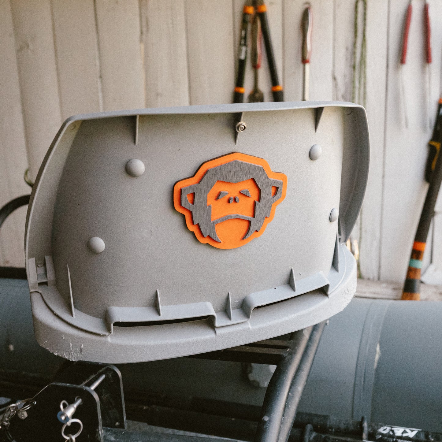 A gray hard hat with an orange and black monkey face logo rests on equipment in a workshop, featuring the El Mono Hook Pad secured with industrial-strength adhesive, as tools hang on a wooden wall in the background. - View 3