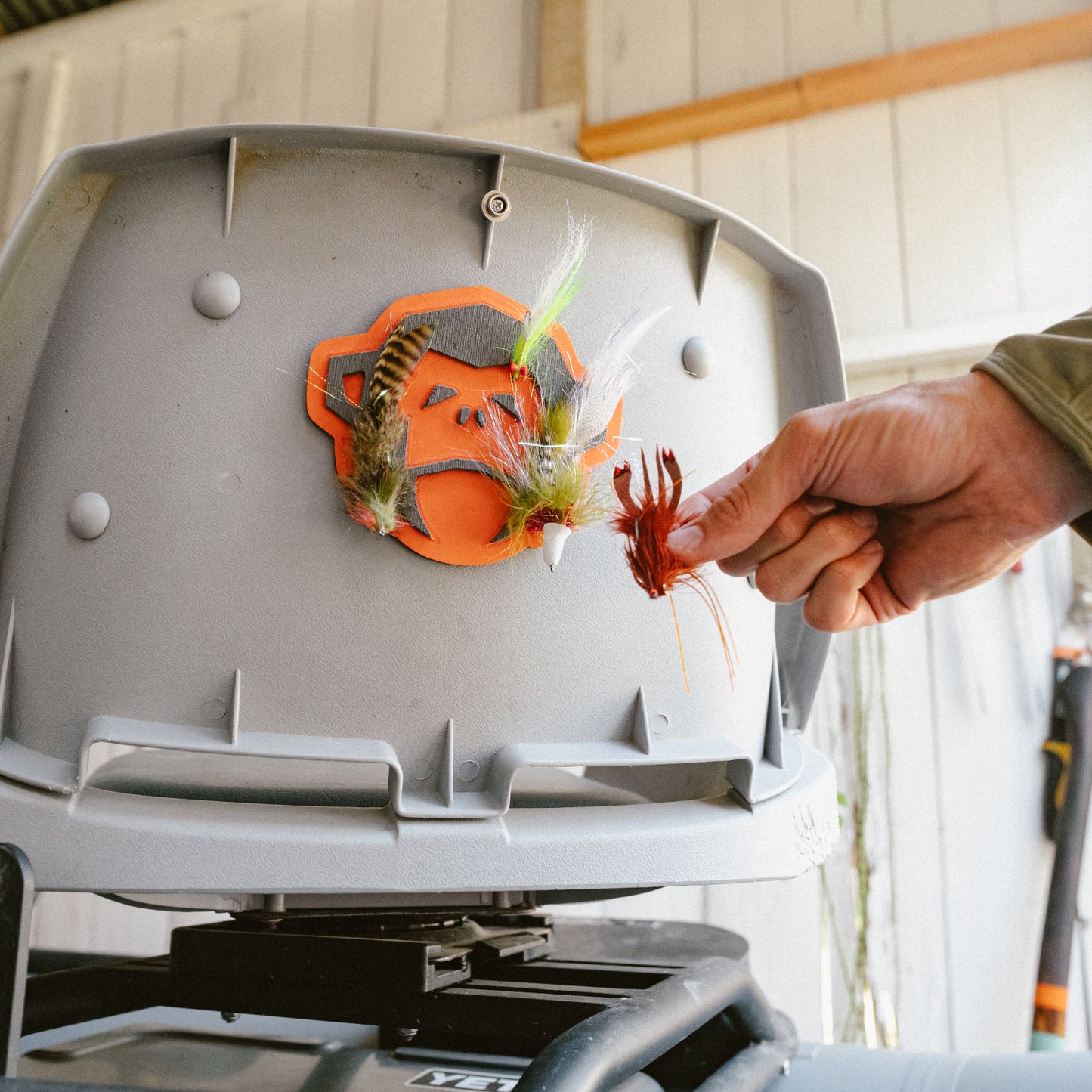 A hand places colorful fishing flies onto the El Mono Hook Pad, a high-density foam fly holder attached to a gray container inside a shed or garage. - View 2