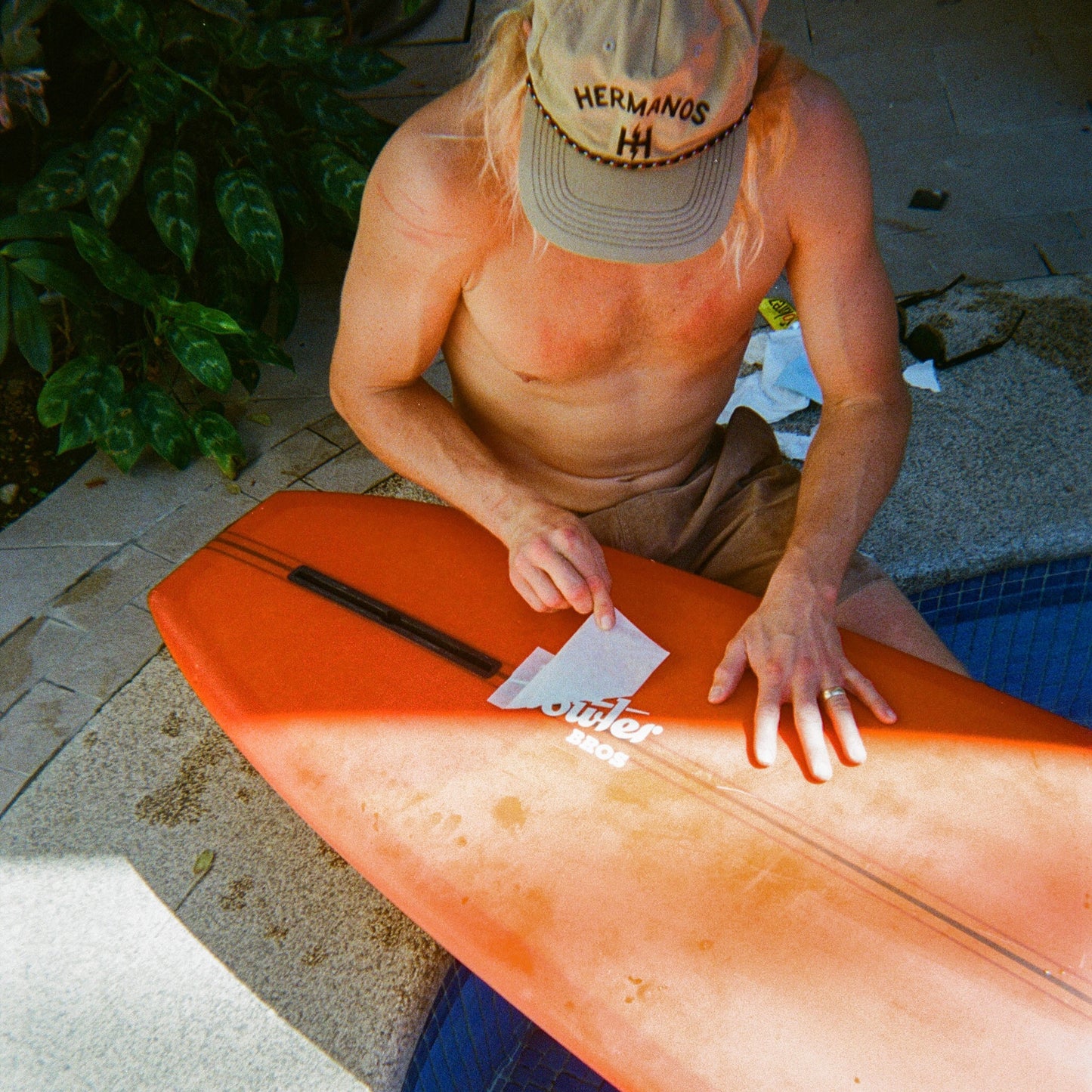 A shirtless person wearing a beige HERMANOS hat sits near a pool, waxing an orange surfboard decorated with Howler Brothers stickers. Lush green plants are nearby, and sunlight casts shadows on the ground. - View 2