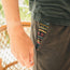 A person’s hand rests by their side, wearing dark French terry sweatpants with a colorful patterned fabric peeking from the pocket and a dark shirt. In the background, blurred greenery and part of a woven chair can be seen.