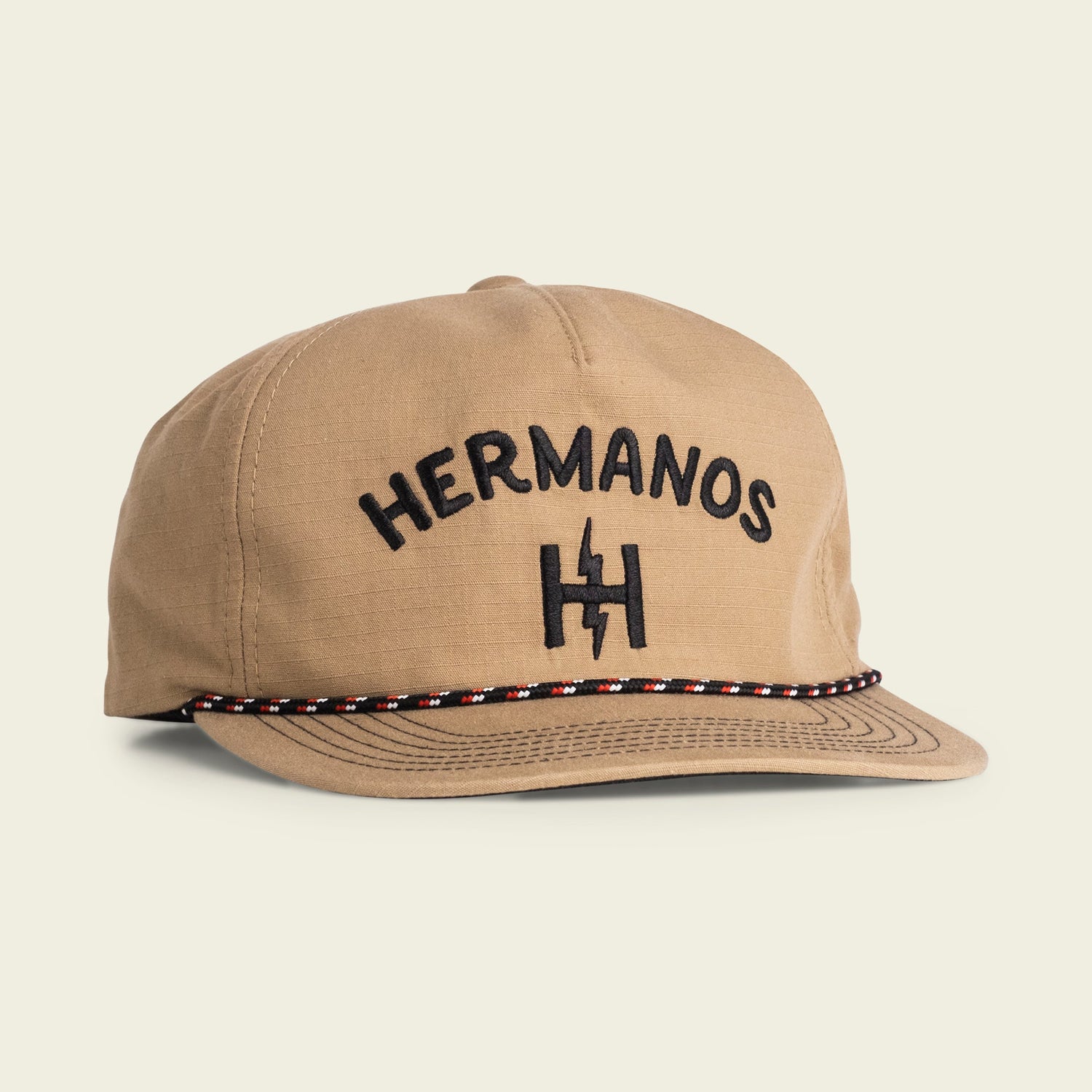 A tan baseball cap with HERMANOS and a stylized H logo embroidered in black on the front. This soft crown, unstructured snapback features a decorative black, white, and red braided band on the brim and an adjustable snapback closure for comfort.