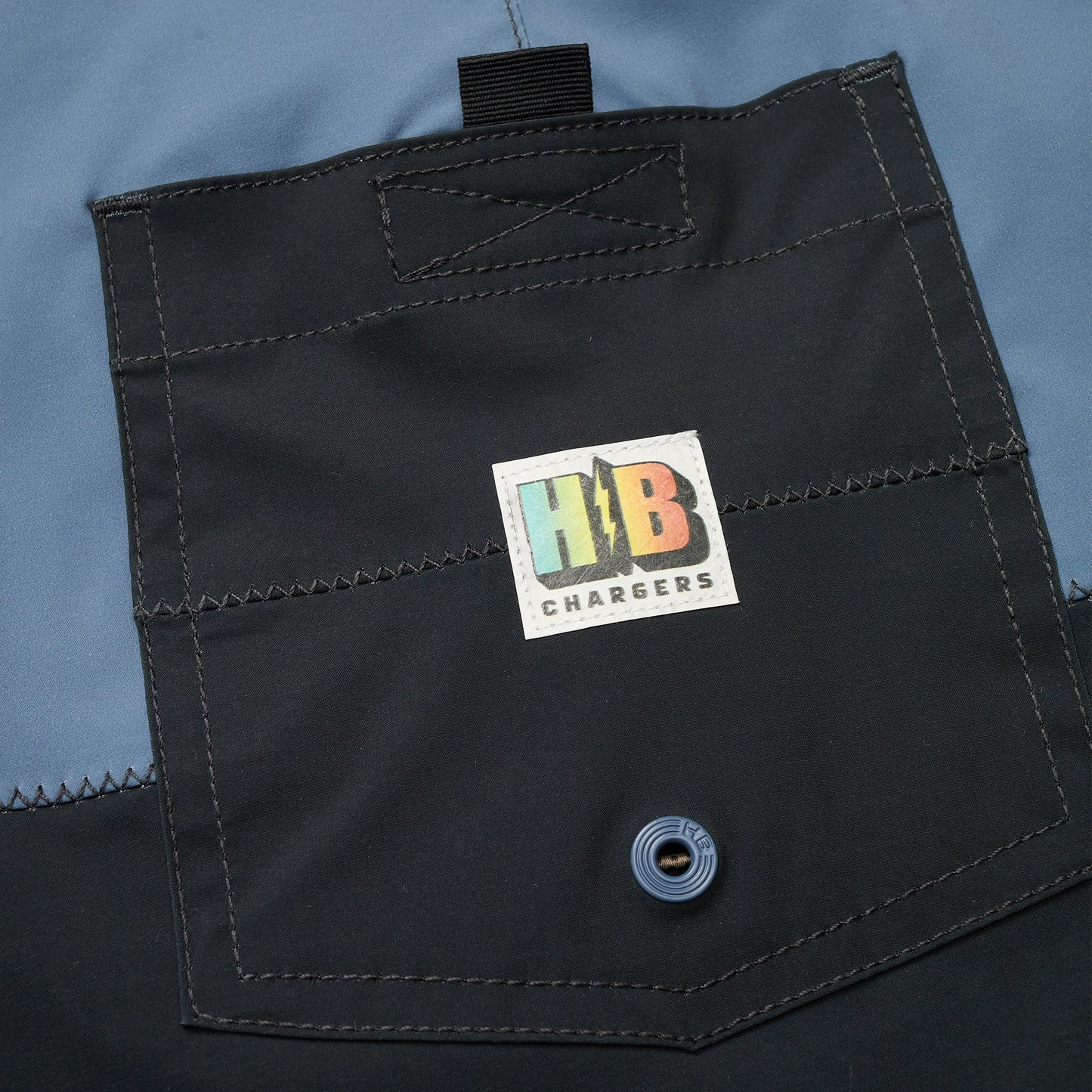 Close-up of a dark fabric pocket with visible stitching, a blue snap button, and a colorful rectangular H1B CHARGERS logo in the center of the flap—perfect for Buchannon Boardshorts with quick drying performance. - View 3