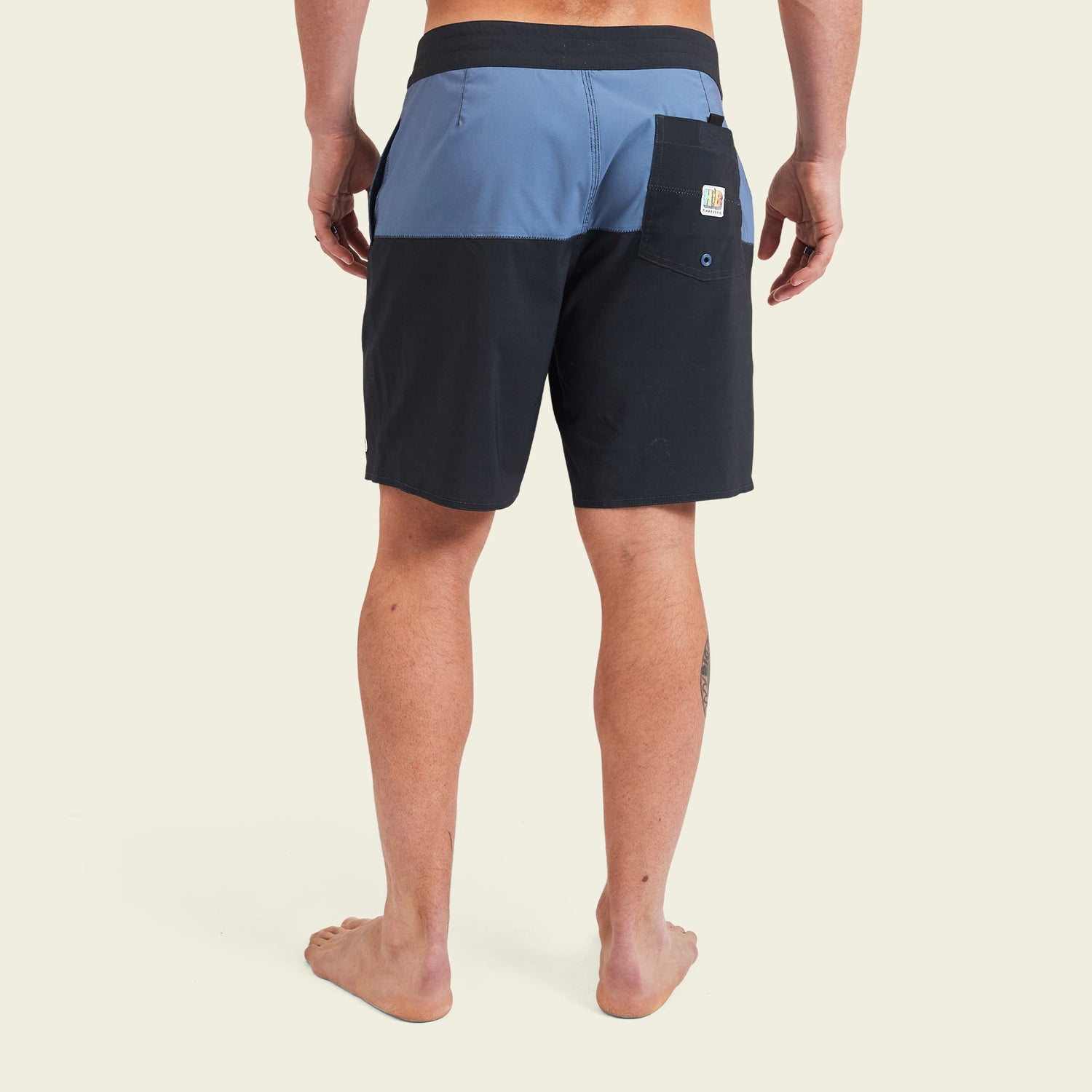 A barefoot person stands facing away, wearing black and blue color-blocked Buchannon Boardshorts with a back pocket and a small patch logo above the pocket, perfect quick drying surf shorts against a plain light background. - View 5