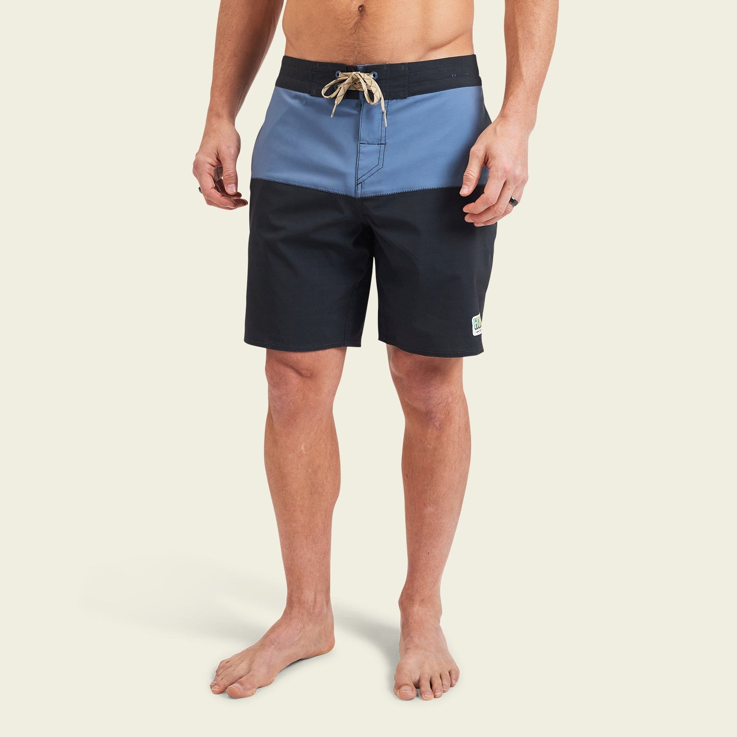 A person wearing Buchannon Boardshorts—the quick drying, two-tone surf shorts with a blue upper section, black lower section, drawstring waist, and a small patch near the left hem—stands barefoot against a plain background. - View 4