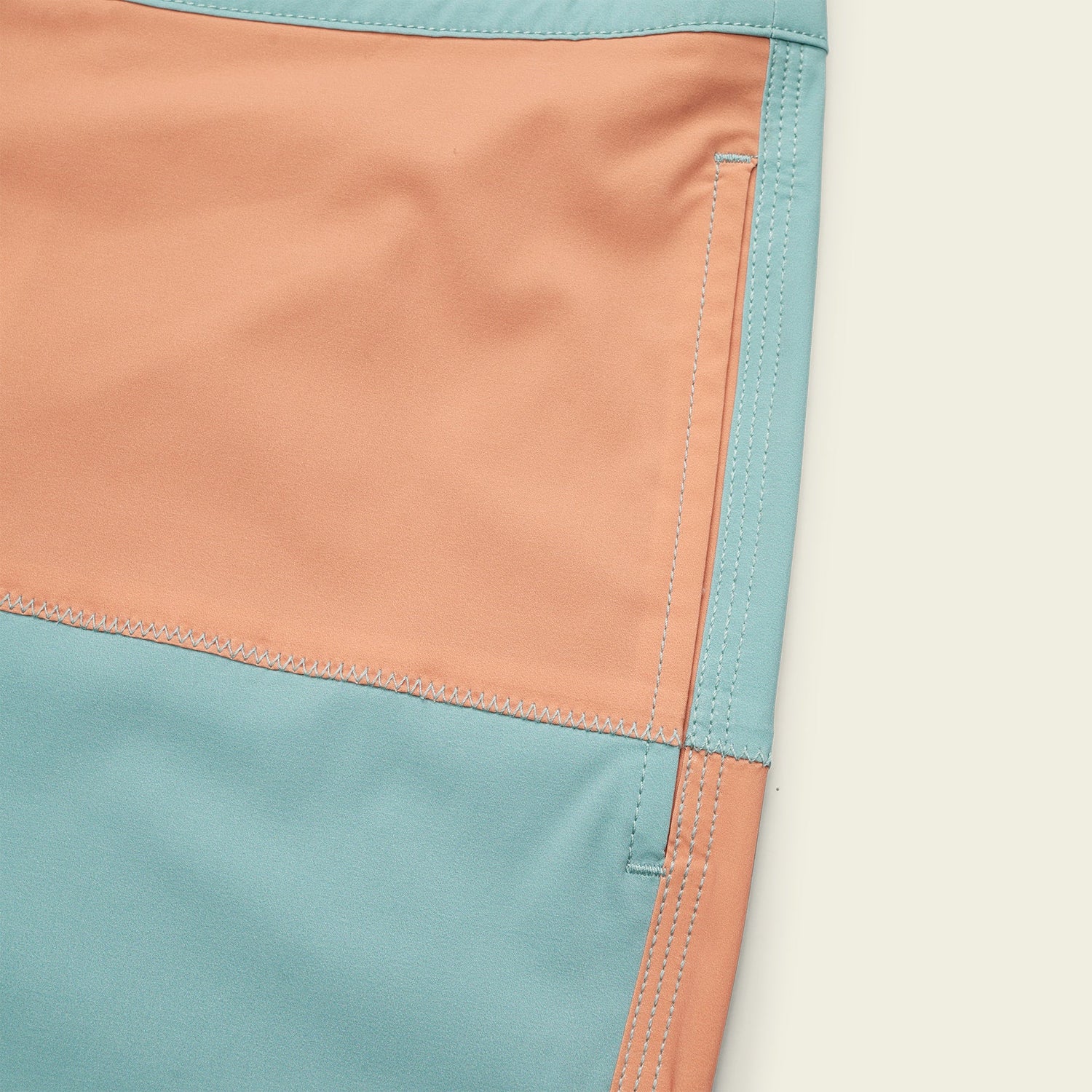 Close-up of quick drying surf shorts made from peach and light blue fabric panels with visible stitching and a vertical pocket. The image highlights the seam details and color blocking design of these boardshorts. - View 3