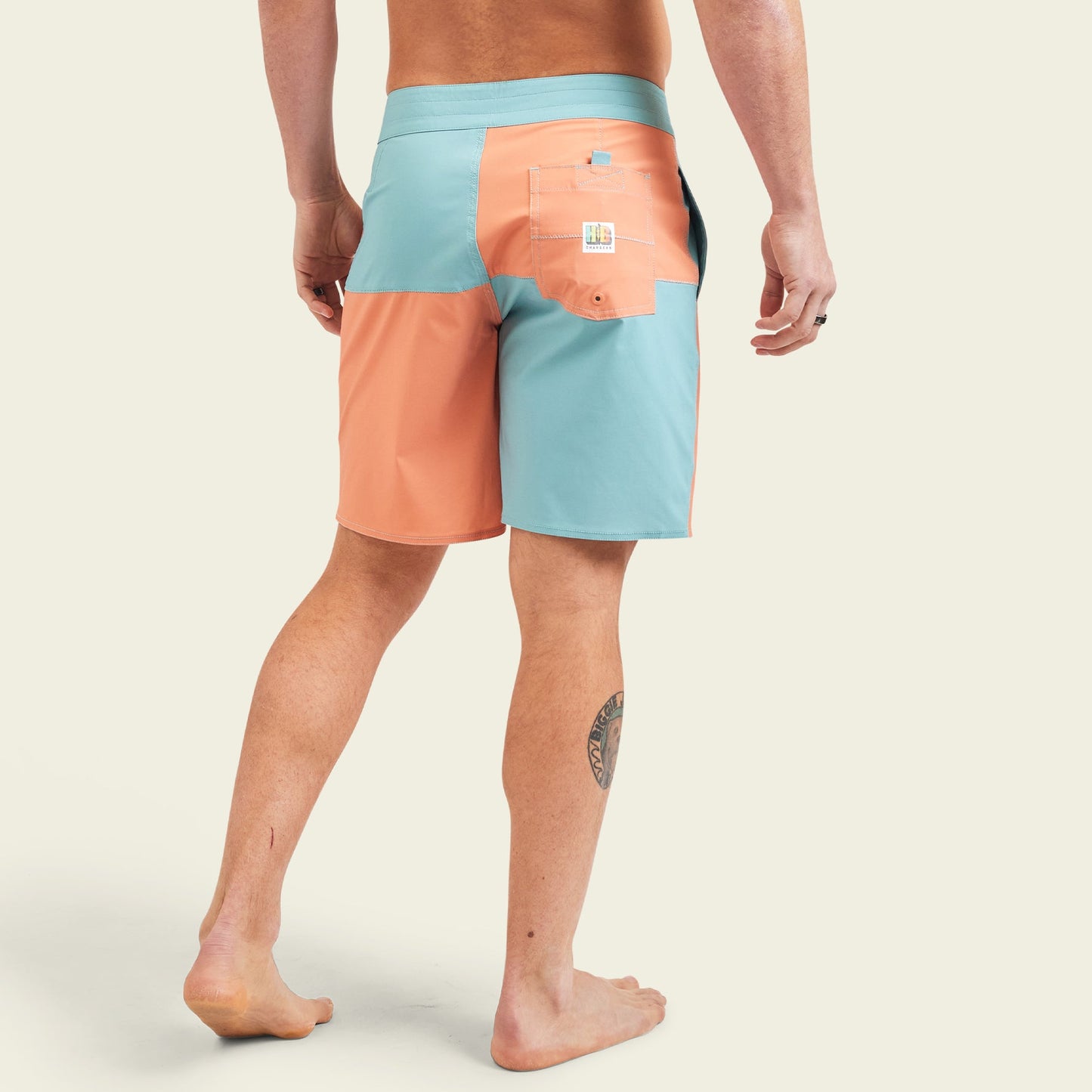 A person stands barefoot, showing the back of their legs and wearing quick drying boardshorts in peach and light blue with a pocket and logo on the right side. A tattoo is visible on the back of their left calf. - View 5