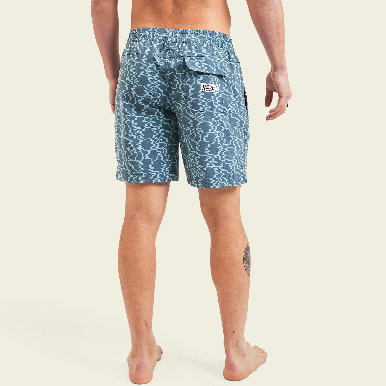 A person stands barefoot with their back to the camera, wearing blue patterned beach shorts with an elastic waistband, a back pocket, and a small white patch. The background is plain and light-colored. - View 4
