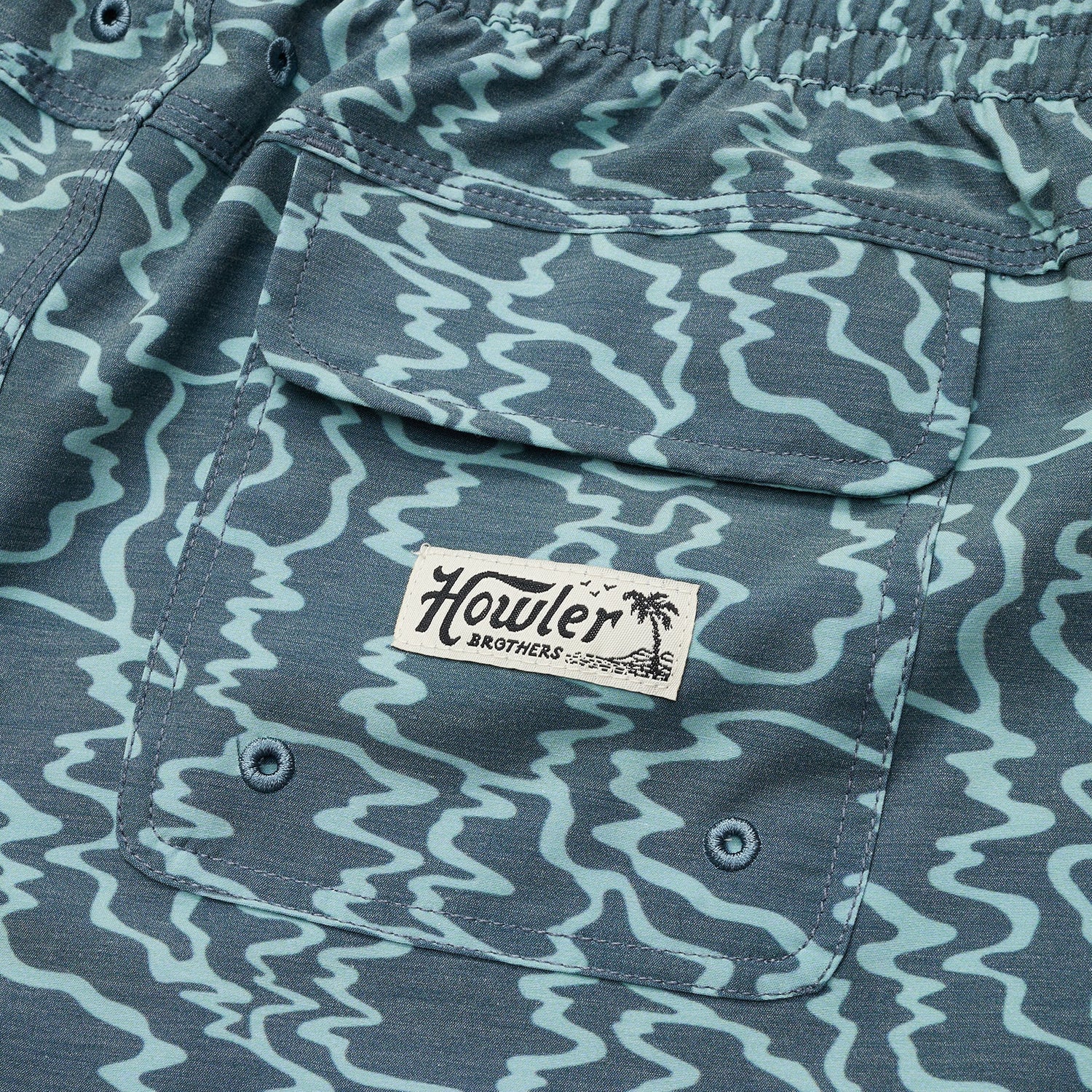 Close-up of blue beach shorts with a wavy light blue pattern, featuring a flap pocket with the Howler Brothers brand label and palm tree logo. The elastic waistband is visible at the top. - View 5