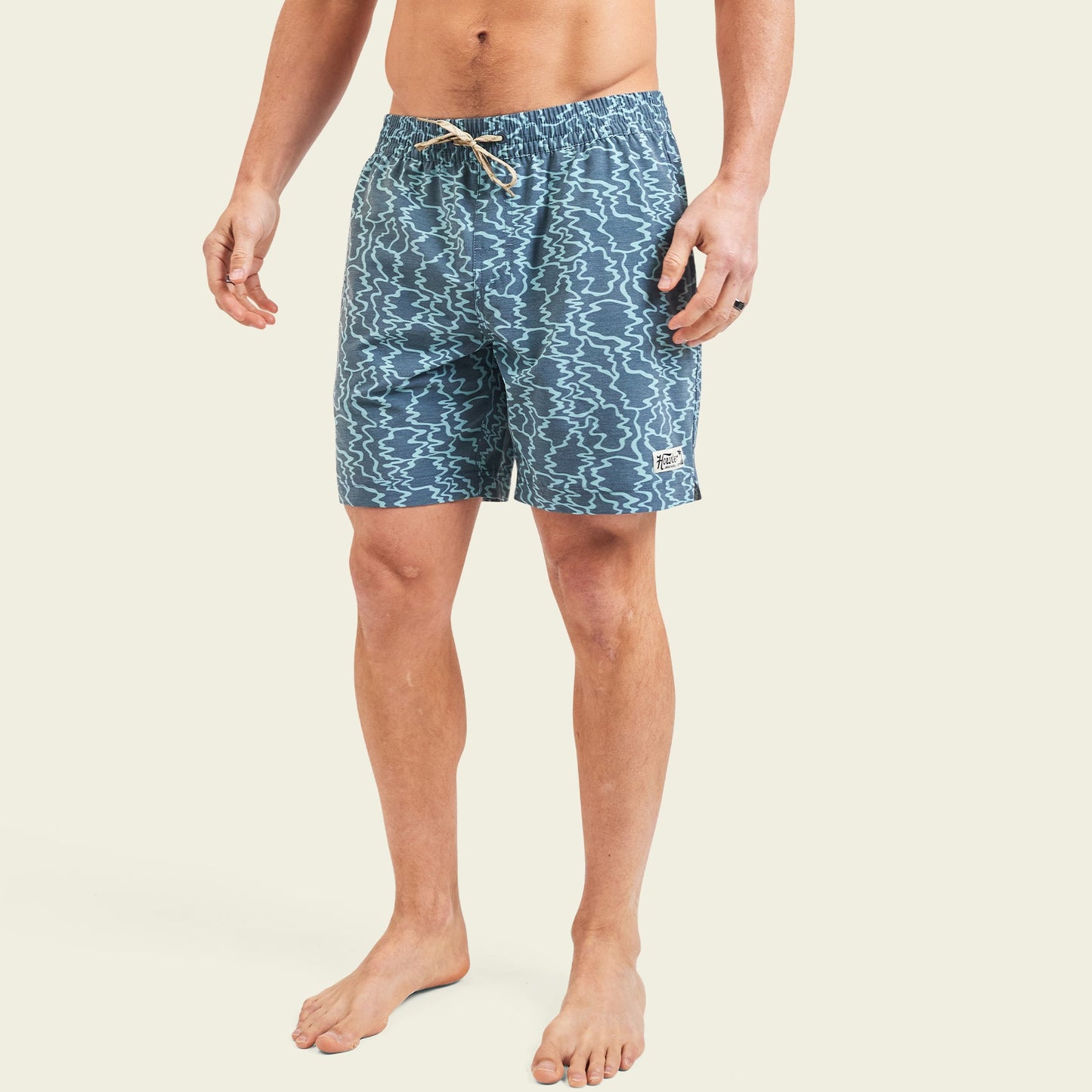 A person stands barefoot wearing blue patterned beach shorts with an elastic waistband and drawstring, set against a plain light background. - View 3